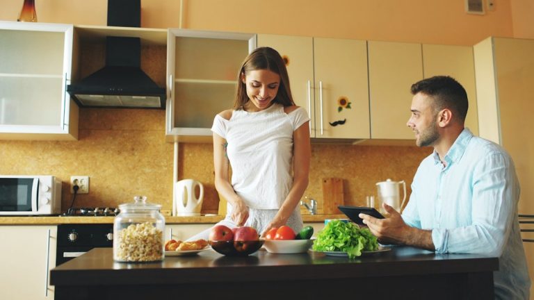 Attractive couple chatting in the kitchen early morning. Handsome man using tablet while his girlfriend cooking breakfast at home